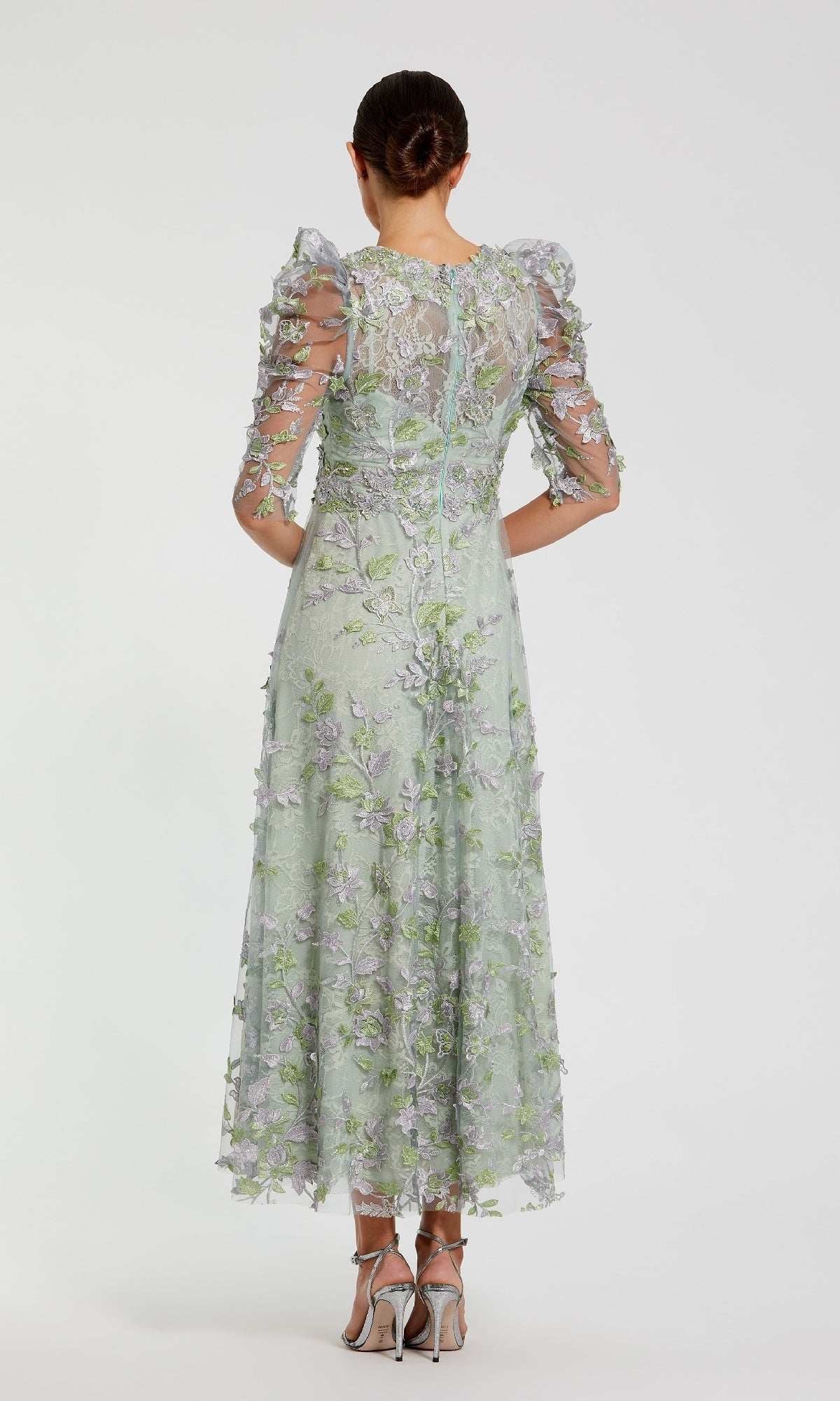 A woman stands facing away, wearing the Mac Duggal 68193 Tea-Length Lace Formal Dress in light green with sheer sleeves and embellishments, paired with silver high-heeled sandals.